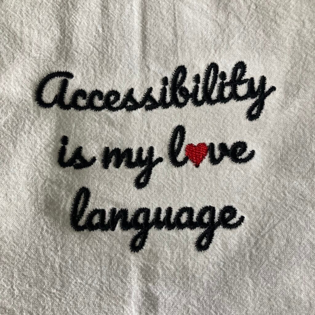 access_square Black script text saying "accessibility is my love language" on a white tea towel, with a red heart for the o in love